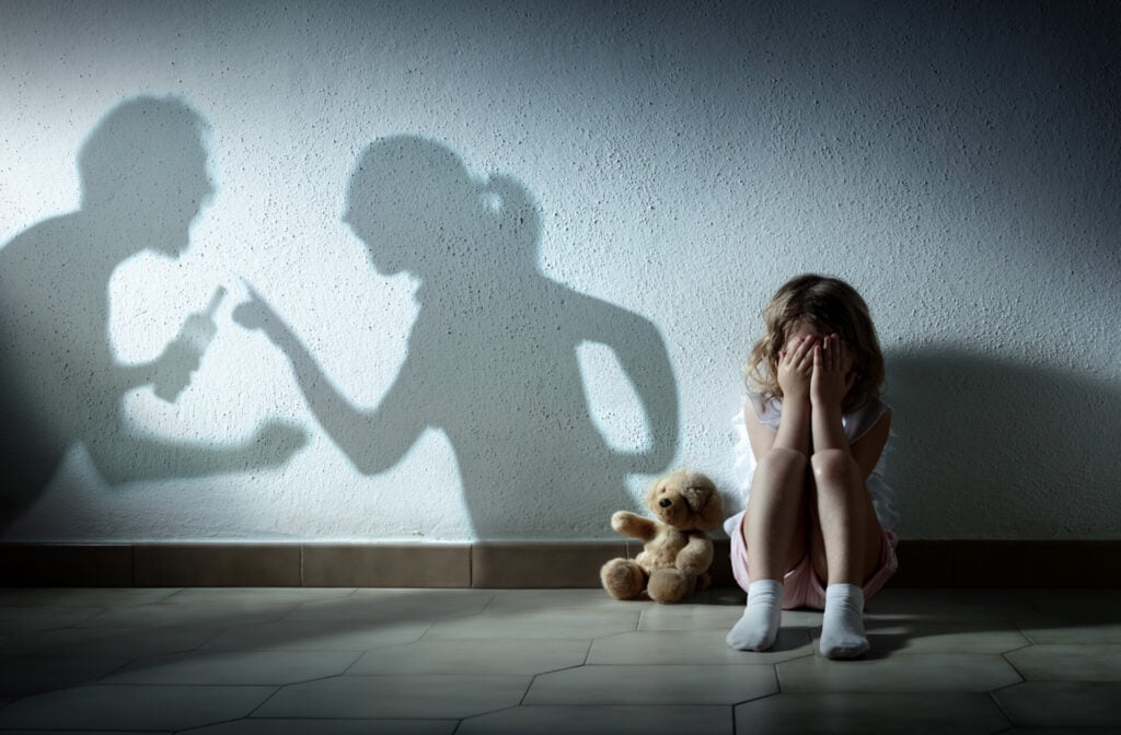 A little girl sat on the floor with face in her hands while there is the shadow of her parents arguing on the wall