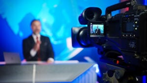 Professional TV Camera Standing in Live News Studio with Anchor seen in Small Display. Unfocused TV Broadcasting Channel with Presenter, Newscaster Talking. Mock-up Television Channel Newsroom Set